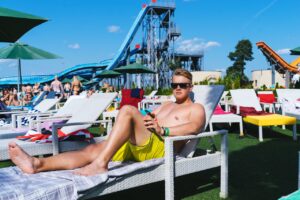 A man enjoys sunbathing by the pool at a lively water park resort on a sunny day.